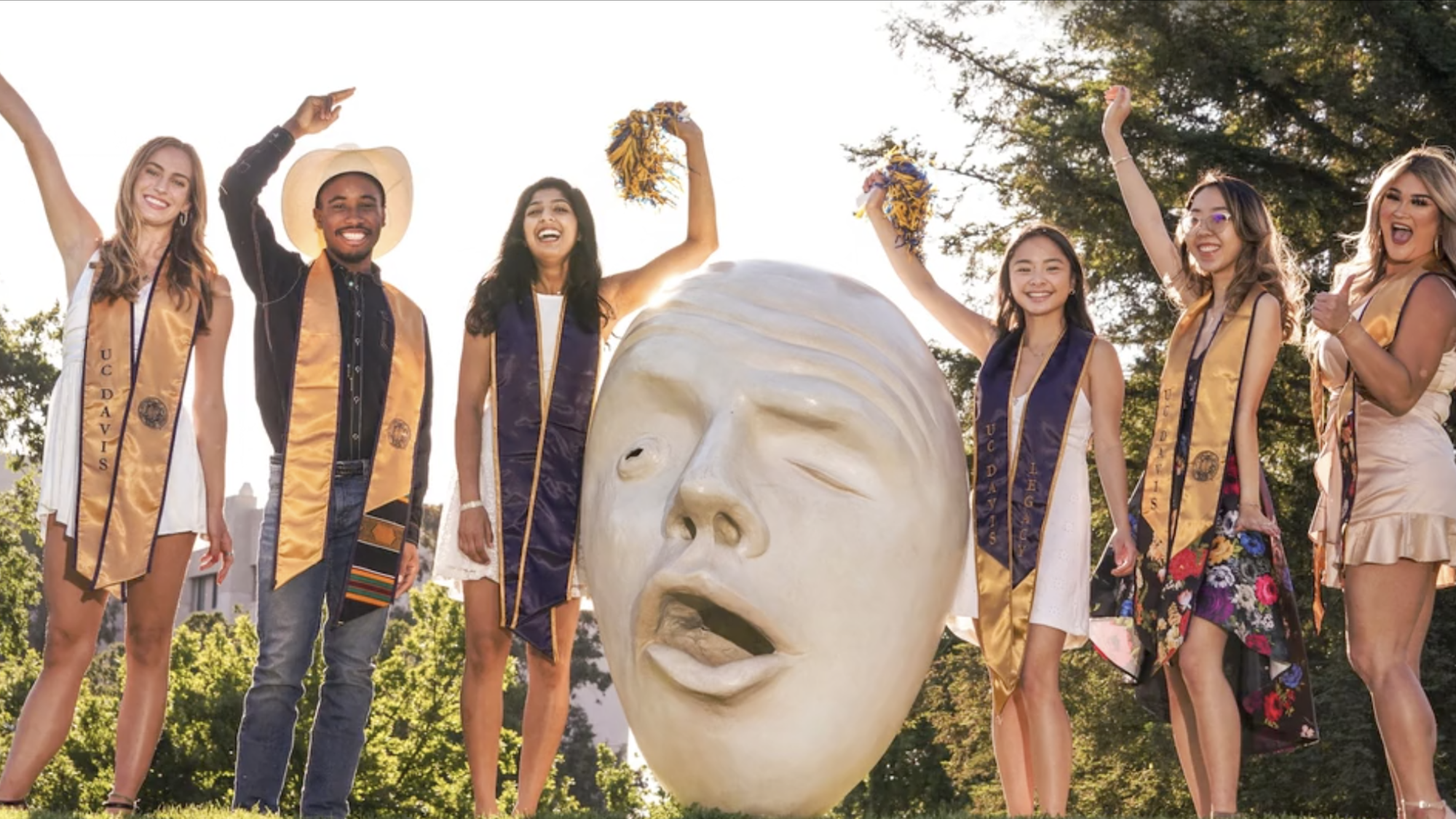 six students in graduation hats and sashes pose near an Egghead statue at ֱ