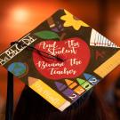 A grad cap that reads, "And the student became the teacher," in white cursive within a bright red apple, surrounded by teaching equipment like rulers, crayons and a pencil that reads, "Ms. Murillo." The photo was taken at ֱ' School of Education Commencement graduation ceremony in 2023.