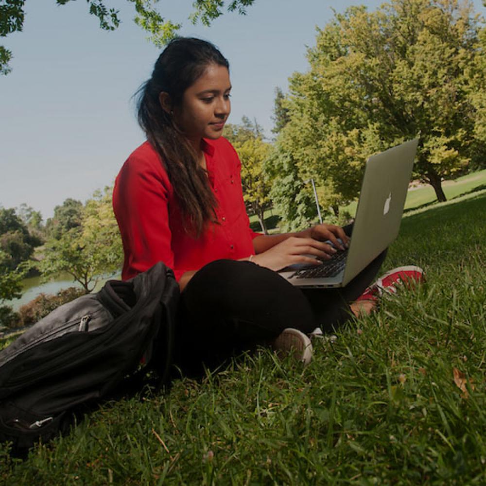 A female student in red reads from her laptop on the ֱ quad