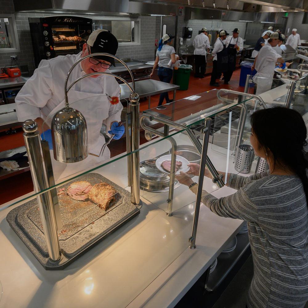 A dining commons employee serves a cut of meat to a ֱ student