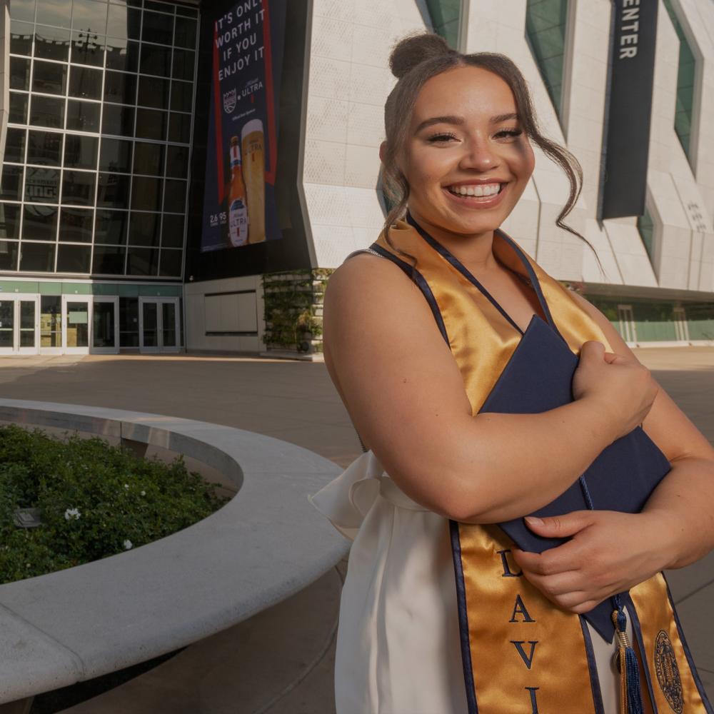 A ֱ student standing outside of the Golden 1 center holdint their grad regalia